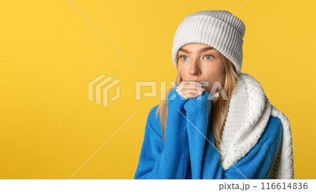 A young woman wearing a blue sweater and white beanie stands pensively against a bold yellow background. Her white scarf is draped over her shoulder while she appears lost in thought. 116614836