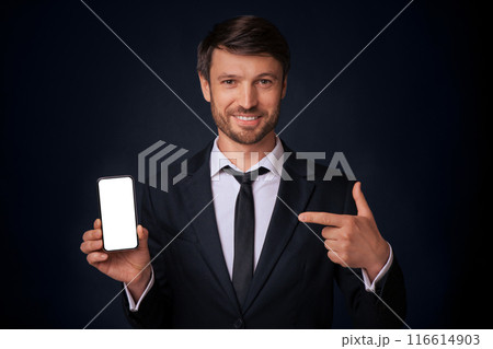 Business App. Cheerful Man Pointing Finger At Blank Mobile Phone Screen Standing On Black Studio Background. Mockup Business App. Cheerful Man Pointing Finger At Blank Mobile Phone Screen Standing On Black Studio Background. Mockup 116614903