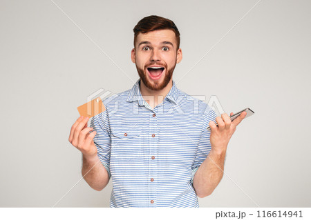 Easy payments. Happy young man holding credit card and cellphone in his hands, isolated on grey studio background, free space Easy payments. Happy young man holding credit card and cellphone in his hands, isolated on grey studio background, free space 116614941
