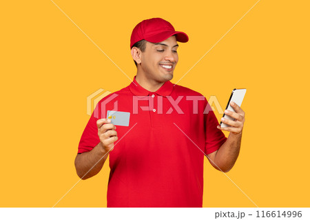 A cheerful delivery worker in a red uniform and cap is seen holding a credit card and a smartphone. He appears engaged as he looks at the smartphone screen, standing against a vivid yellow background. 116614996
