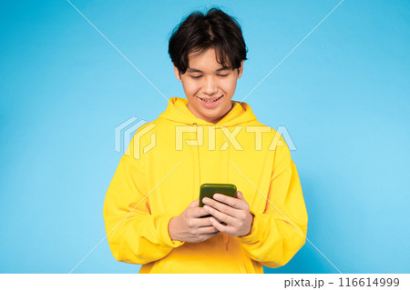 Asian guy wearing a yellow hoodie is focused on his cell phone screen, engrossed in whatever he is viewing or reading. 116614999
