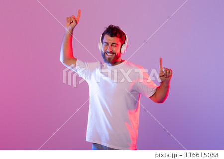 A cheerful man wearing wireless headphones is dancing and enjoying music. The background features vibrant, colorful studio lighting, enhancing the lively atmosphere. A cheerful man wearing wireless headphones is dancing and enjoying music. The background features vibrant, colorful studio lighting, enhancing the lively atmosphere. 116615088