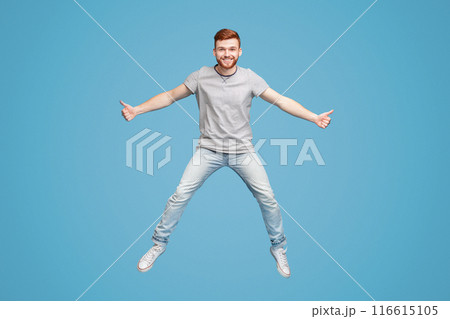 Young millennial redhaired guy jumping in air on blue background, demonstrating thumbs up gesture, full length, studio, empty space 116615105