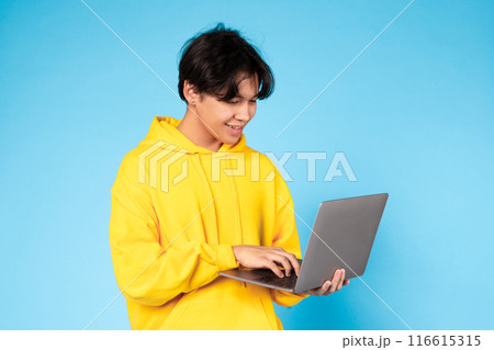 Asian teen guy wearing a yellow hoodie is sitting at a laptop, typing on the keyboard. The laptop screen displays a webpage or document. The person appears focused on their work. 116615315