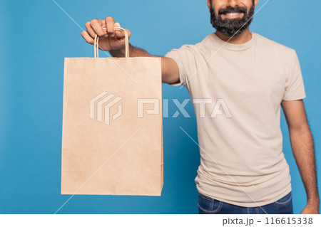 Cropped of cheerful delivery man with a beard is holding a blank brown paper bag in front of a bright blue background 116615338