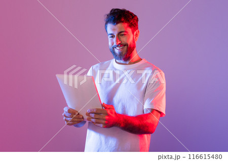 A man wearing a white t-shirt is shown holding a tablet device in his hands. He appears focused on the screen, engaged in technology. 116615480