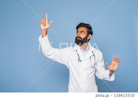 A bearded Indian doctor wearing a white lab coat and stethoscope gestures with his right hand as if pointing towards something, set against a solid blue background. A bearded Indian doctor wearing a white lab coat and stethoscope gestures with his right hand as if pointing towards something, set against a solid blue background. 116615510