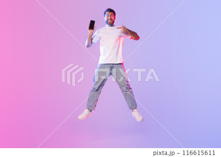 A young man dressed in a white t-shirt and jeans energetically jumps while holding and pointing at his smartphone. The backdrop features an eye-catching gradient of pink and blue A young man dressed in a white t-shirt and jeans energetically jumps while holding and pointing at his smartphone. The backdrop features an eye-catching gradient of pink and blue 116615611