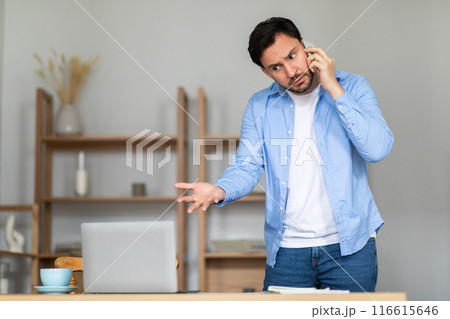 A man stands in his home office, with shelves in the background, gesturing with his hand while talking on a smartphone. He appears puzzled or concerned, possibly discussing an important matter A man stands in his home office, with shelves in the background, gesturing with his hand while talking on a smartphone. He appears puzzled or concerned, possibly discussing an important matter 116615646