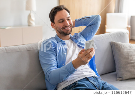 A man is sitting on a couch indoors, engrossed in using his smartphone. He is focused on the screen, tapping and swiping. The room is dimly lit, with soft ambient lighting. A man is sitting on a couch indoors, engrossed in using his smartphone. He is focused on the screen, tapping and swiping. The room is dimly lit, with soft ambient lighting. 116615647