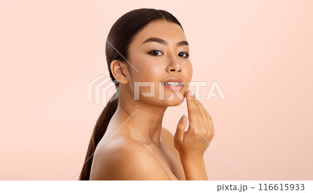 A young Asian woman with brown hair, wearing no makeup, is touching her cheek with her right hand. She is looking to the left, and her expression is relaxed and confident. 116615933