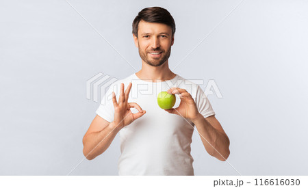 A man in a white t-shirt holds a green apple in one hand and makes an okay sign with the other, smiling at the camera. 116616030