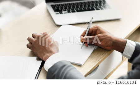 A person's hand writes in a notebook while sitting at a desk, a laptop in the background. 116616131