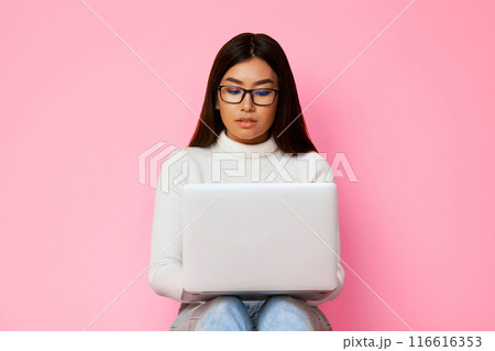 A young Asian woman wearing glasses is sitting and working on a laptop in front of a pink background. She is looking down at the screen and appears to be focused on her work. 116616353