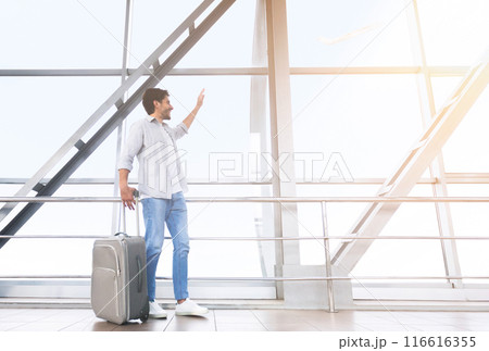 Welcoming New City. Happy Man Traveler Walking With Luggage In Airport, And Waving To Window, Excited About Trip, Free Space Welcoming New City. Happy Man Traveler Walking With Luggage In Airport, And Waving To Window, Excited About Trip, Free Space 116616355
