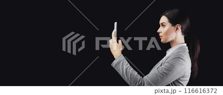 A woman in a grey blazer holds a smartphone in front of her face, looking directly at it. The background is black. 116616372