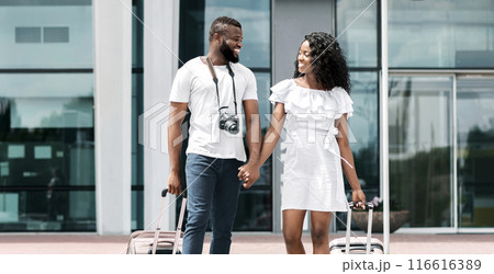 A young couple, dressed in casual summer clothes, are walking through an airport terminal. They are holding hands and pulling luggage behind them. A young couple, dressed in casual summer clothes, are walking through an airport terminal. They are holding hands and pulling luggage behind them. 116616389