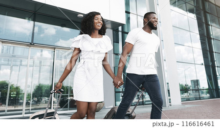 Black couple walks through an airport terminal, pulling luggage behind them. They are both smiling and looking happy, holding hands as they walk towards their destination 116616461