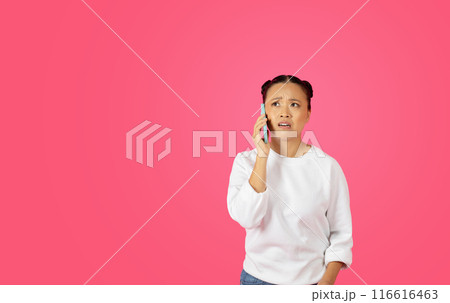 Asian woman with her hair in buns is talking on a cell phone, and she has a confused expression on her face. The background is a solid pink color, copy space Asian woman with her hair in buns is talking on a cell phone, and she has a confused expression on her face. The background is a solid pink color, copy space 116616463