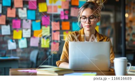 A woman sits at a desk in an office, focusing on her laptop while a wall of colorful sticky notes serves as a visual reminder. 116616689