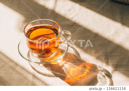 A cup of tea in glass cup and saucer illuminated by warm sunlight on a light table. Horizontal frame, copy space 116621138