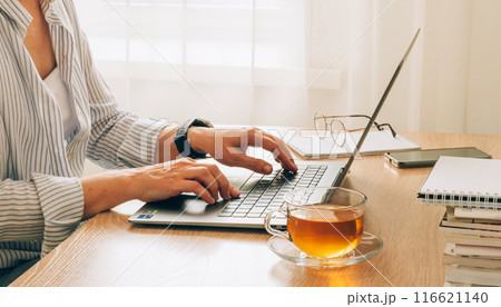Female hands typing on a laptop with cup of tea, glasses and notepads on a wooden table 116621140