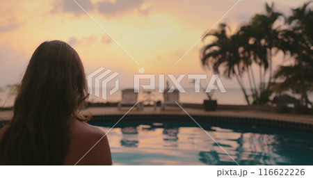 Long hair brunette enjoy bright colorful sunset on tropical island. Orange sky reflected in swimming pool, palm trees and deck chairs. Lifestyle travel on summer holiday vacation. Back view zoom in Long hair brunette enjoy bright colorful sunset on tropical island. Orange sky reflected in swimming pool, palm trees and deck chairs. Lifestyle travel on summer holiday vacation. Back view zoom in 116622226