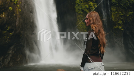 A woman stands in front of a majestic Bali waterfall, surrounded by lush greenery. The water cascades down, creating a breathtaking natural spectacle. 116622255