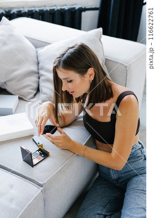 Young woman is sitting on the couch and looking at her makeup. She is holding a compact mirror and a bottle of cosmetics Young woman is sitting on the couch and looking at her makeup. She is holding a compact mirror and a bottle of cosmetics 116622761