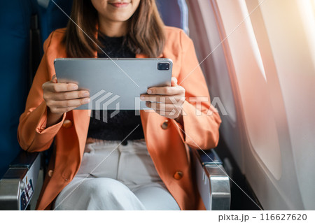 Close-up of a woman in an airplane seat using a tablet, dressed in an orange blazer and white pants, with sunlight streaming in. 116627620