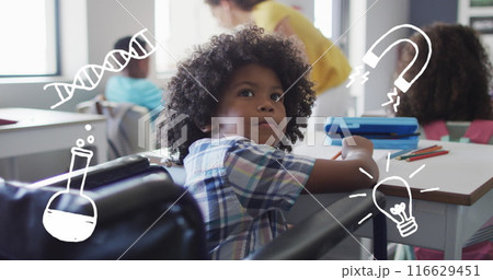 Image of science icons over happy biracial schoolboy in wheelchair at desk in class 116629451
