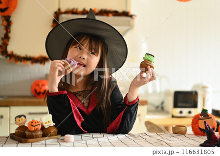 Cute little girl in witch costumes standing behind kitchen table with Halloween desserts Cute little girl in witch costumes standing behind kitchen table with Halloween desserts 116631805