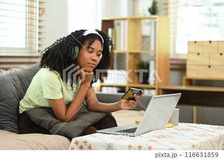 Young African American woman wearing headphone using mobile phone in living room Young African American woman wearing headphone using mobile phone in living room 116631859