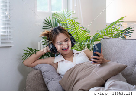 Young Woman Relaxing on Couch with Headphones and Smartphone in Modern Living Room Young Woman Relaxing on Couch with Headphones and Smartphone in Modern Living Room 116633111