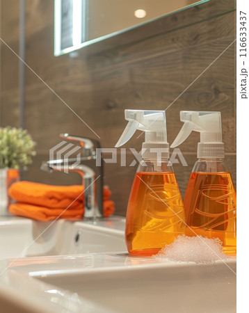 Close-up of a modern bathroom sink with two bottles of orange cleaning solution and orange towels in the background. The scene is clean, organized, and well-lit, showcasing hygiene and cleanliness. Close-up of a modern bathroom sink with two bottles of orange cleaning solution and orange towels in the background. The scene is clean, organized, and well-lit, showcasing hygiene and cleanliness. 116633437