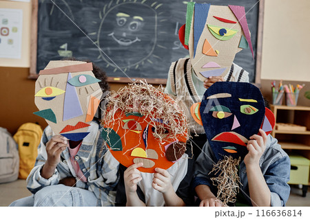 Shot of unrecognizable kids hiding behind cardboard face mask crafts in abstract style in classroom with blackboard 116636814