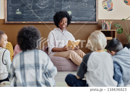 Female teacher of Black ethnicity in bean bag chair reading fairy tale aloud to attentive children sharing thoughts about story sitting around in classroom 116636823