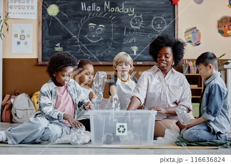Multiethnic group of children and female teacher in ecology club sorting waste putting plastic bottles in recycling bin in classroom 116636824