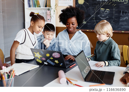 Diverse group of primary learners around female teacher of Black ethnicity explaining new information using laptop sitting at desk during elective lesson 116636890