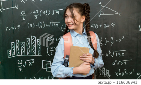 A high school schoolgirl stands near the blackboard with a textbook in her hands, portrait of a teenage girl at school A high school schoolgirl stands near the blackboard with a textbook in her hands, portrait of a teenage girl at school 116636943