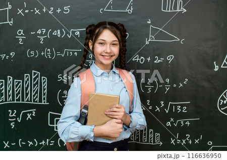 A high school schoolgirl stands near the blackboard with a textbook in her hands, portrait of a teenage girl at school A high school schoolgirl stands near the blackboard with a textbook in her hands, portrait of a teenage girl at school 116636950