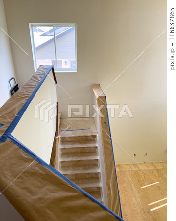 A well-lit stairway prepared for renovation, with protective paper and tape covering the banister. The steps are bare, indicating a work-in-progress state. The window in the background allows natural A well-lit stairway prepared for renovation, with protective paper and tape covering the banister. The steps are bare, indicating a work-in-progress state. The window in the background allows natural 116637865
