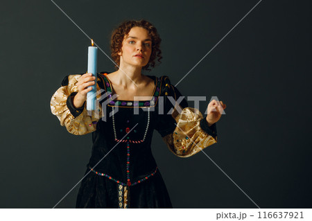 Portrait of a young adult woman dressed in a medieval dress holds a candle on dark background 116637921