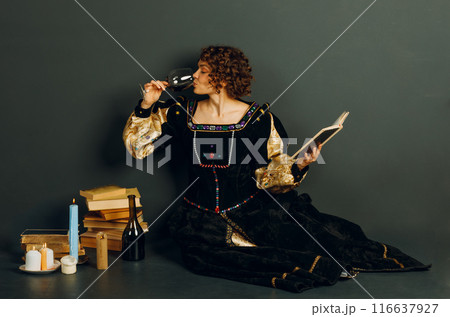 Young adult woman dressed in a medieval dress sitting near ancient books and candles with bottle of red wine and drinking a glass of wine 116637927