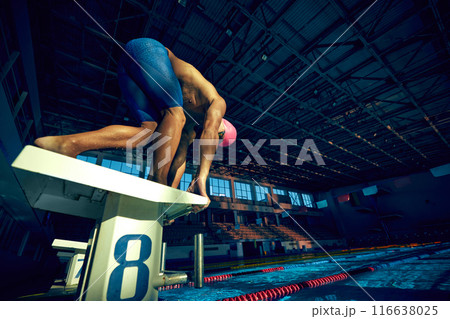 Ready to dive. Low angle view of young muscular man, swimmer, wearing goggles and cap, in low position on starting block in swimming pool 116638025