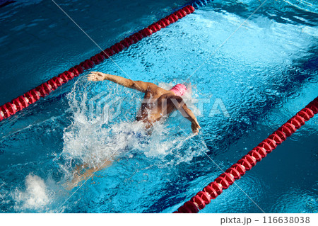 Swimmer in pink cap executes powerful butterfly stroke, his muscular form cutting through blue water, creating dynamic splash between red lane dividers. Swimmer in pink cap executes powerful butterfly stroke, his muscular form cutting through blue water, creating dynamic splash between red lane dividers. 116638038