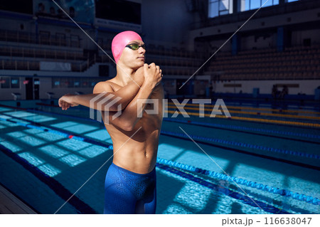 Young man with athletic body, swimmer wearing goggles and pink cap, warming up before swim against blurred background of pool. 116638047