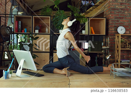 Woman, employee sitting on desk in headphones and practicing yoga and stretching exercises. Healthy lifestyle and contemporary workspace 116638583