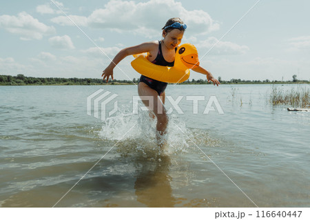 Smiling girl with an inflatable yellow duck running through the lake on a sunny summer day. She enjoys playing and splashing in the water Smiling girl with an inflatable yellow duck running through the lake on a sunny summer day. She enjoys playing and splashing in the water 116640447