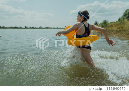 Smiling girl running in the lake with inflatable yellow toy, creating splashes A happy child with an inflatable yellow duck is splashing in the lake on a bright summer day 116640452